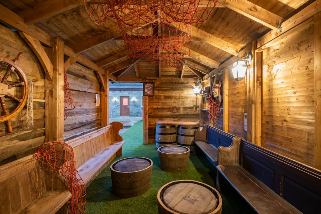 inside a pirate themed shed with a wheel on the wall and barrels on the floor.