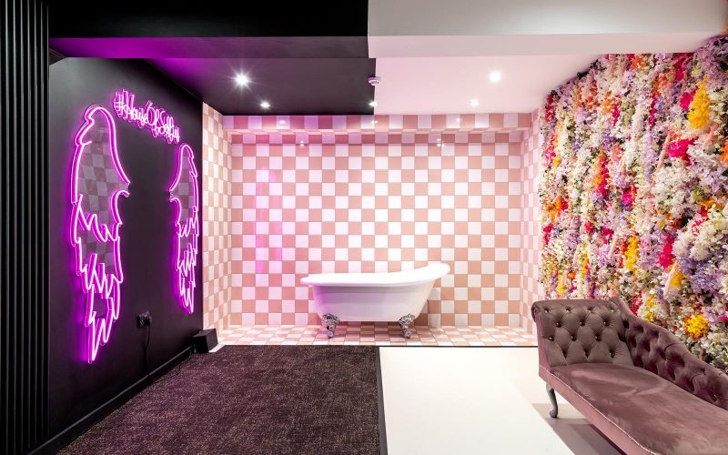 selfie wall with neon pink wings on and a checkerboard background with a bathtub.