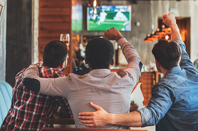 Three men watching football on TV in sport bar, back view.