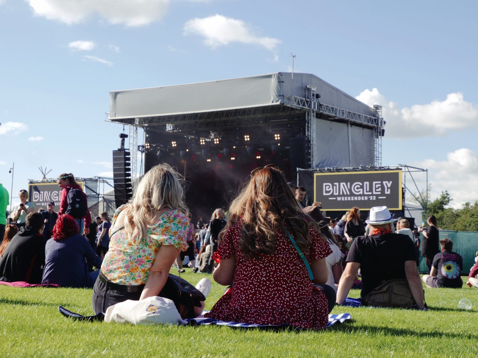 Two women sat on grass watching live music. 