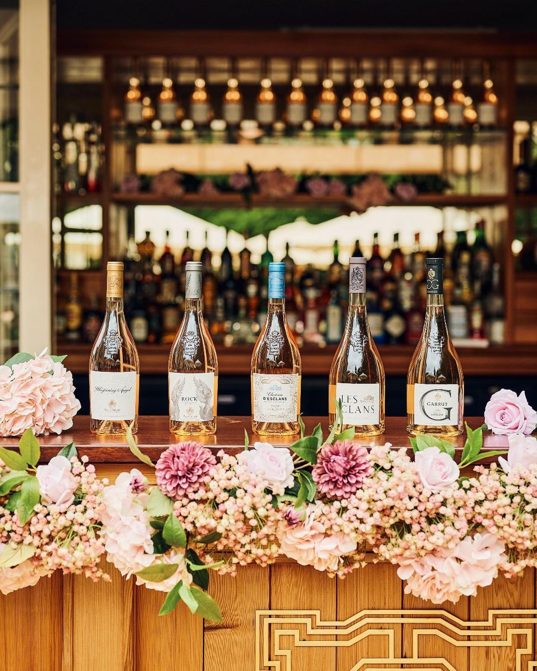 Bottles of rosé lines up on a brown bar covered in pink flowers. 