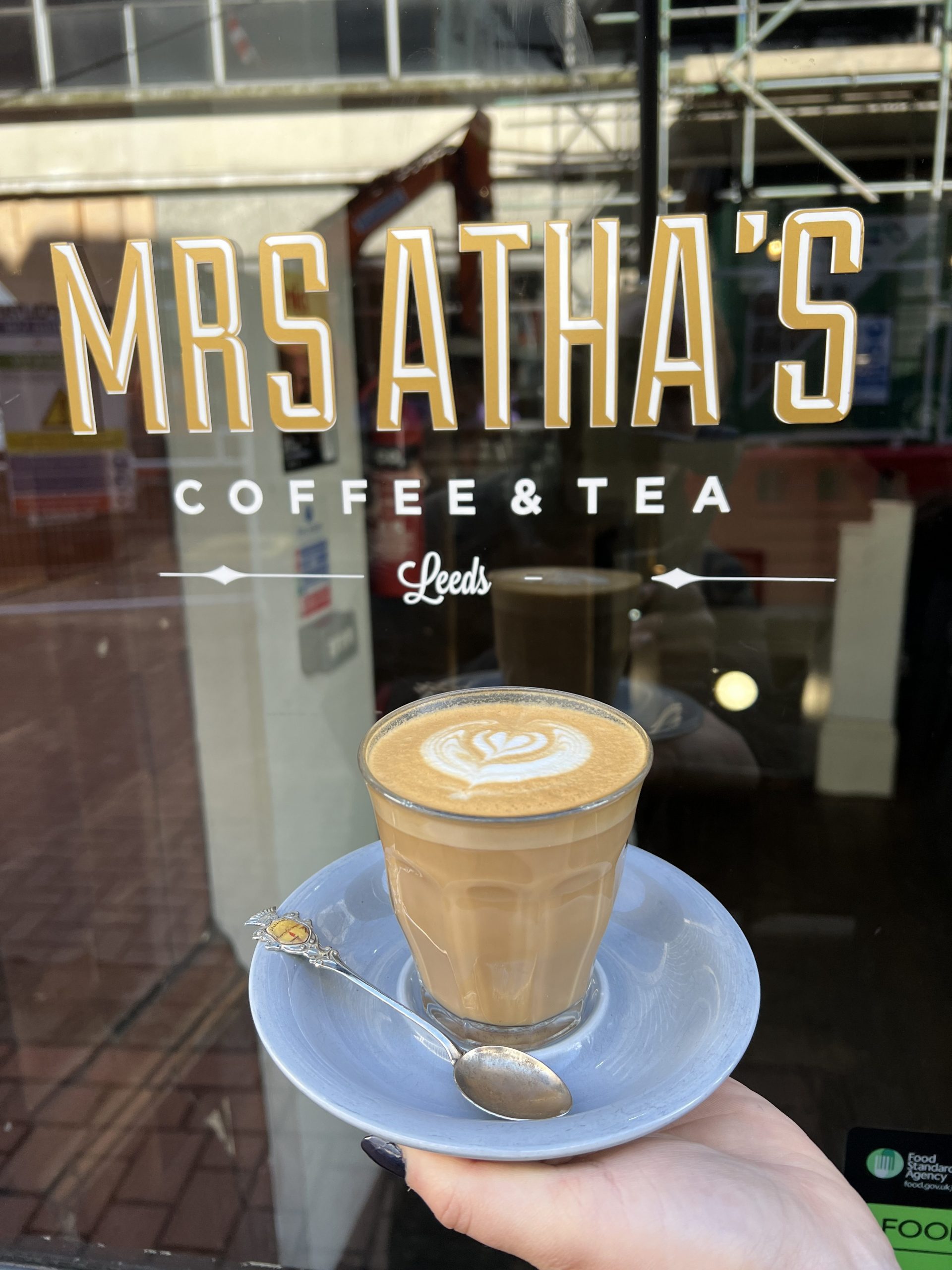 a latte held outside a sign reading 'Mrs Athas Coffee & Tea, Leeds'