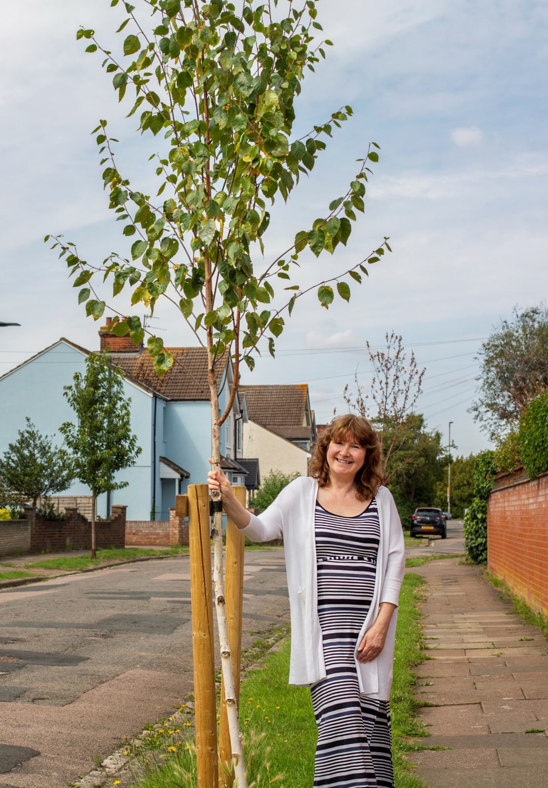 person stood next to a tree with a blue house in the background.