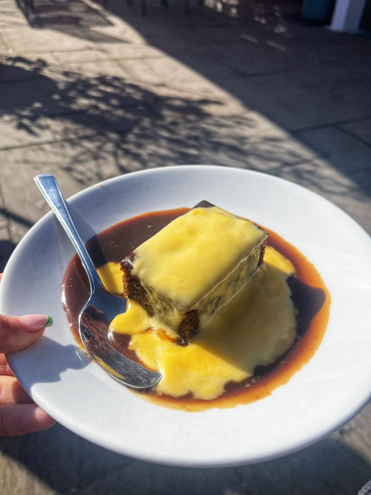 a bowl of sticky toffee pudding with custard on top and a spoon on the left.