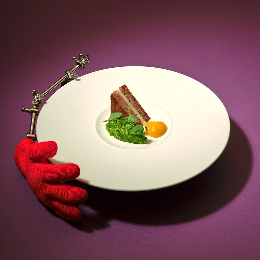 Smoked Ham Hough “Sandwich”, Garden Pea Pesto, Egg Yolk Jam with a red hand holding the plate.
