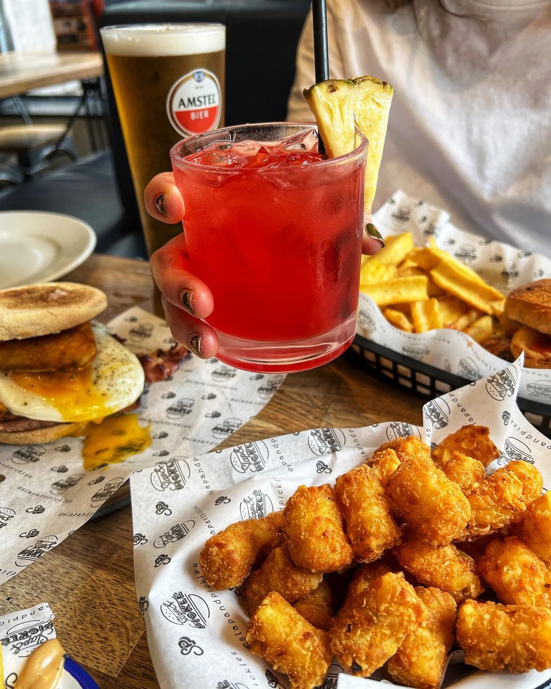 tater tots with a person holding a red cocktail with a pineapple stick in it over the top. There is a burger and chips in the background.