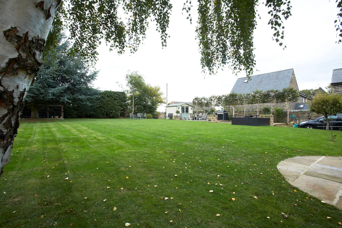 a garden with grass and circle of patio area. There is a car and a shed in the distance.