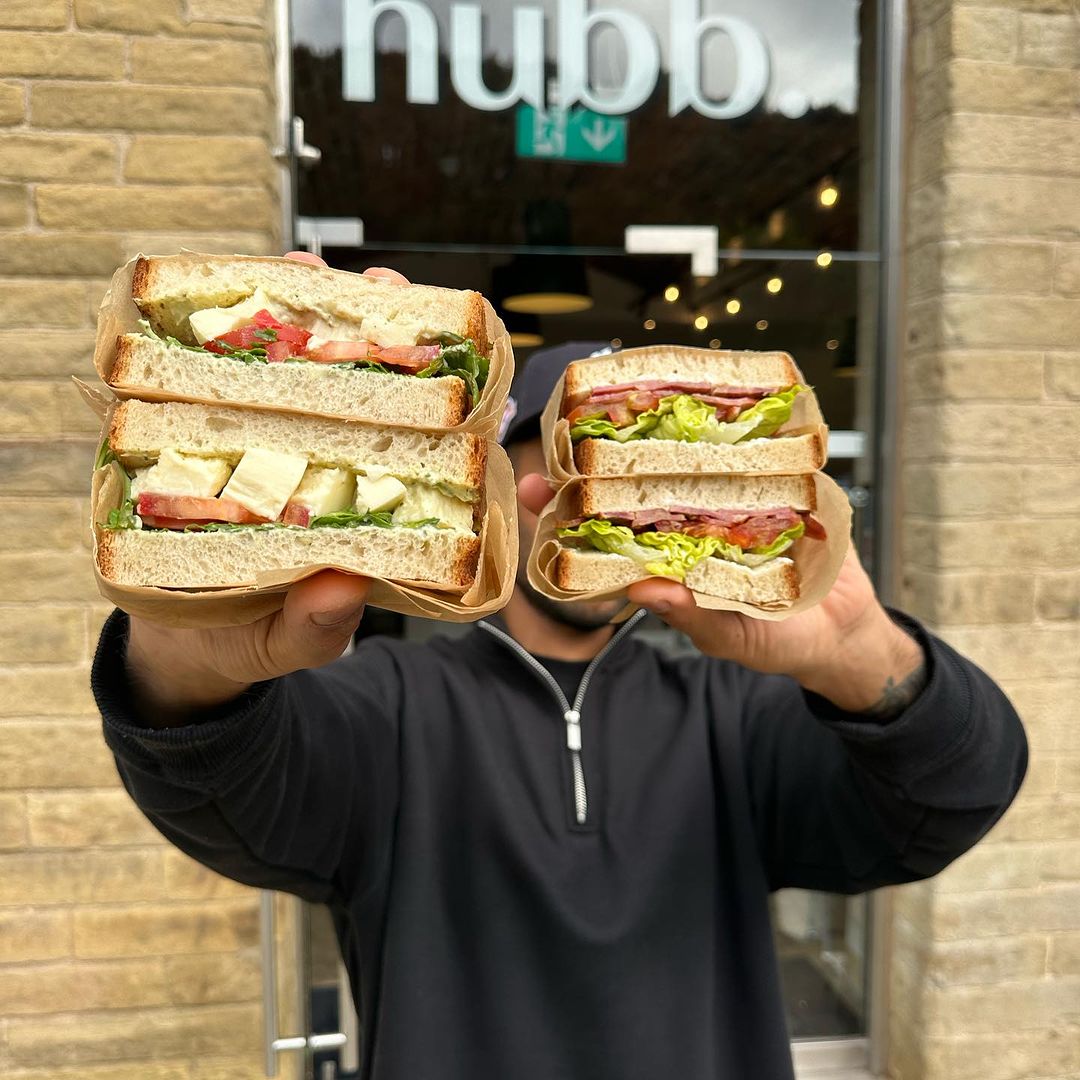 Two sandwiches being held up outside hubb coffee bar. 